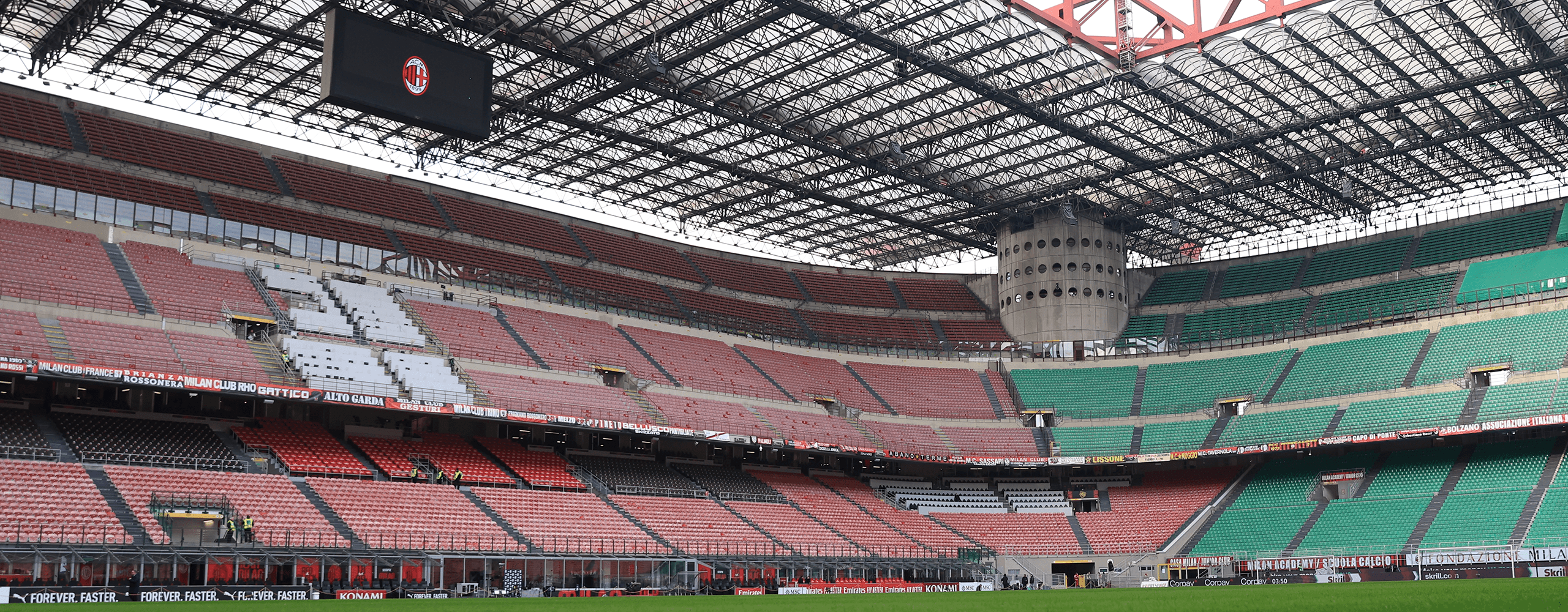 Vista interna di San Siro vuoto con tribune rosse e verdi, struttura di copertura e maxi schermo con logo Milan